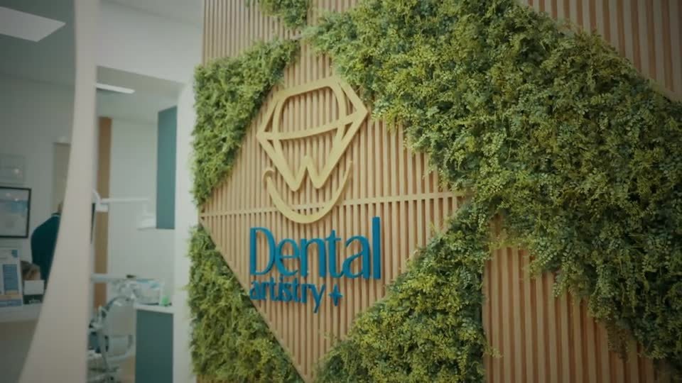 The practice reception: the Dental Artistry mark in gold on a slatted wood wall framed by a living green wall, with a treatment room visible beyond