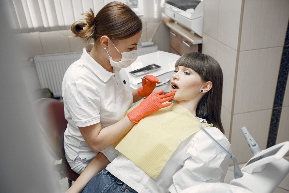 A dentist in a white polo shirt and orange gloves examines a young woman's teeth with a dental mirror while she sits in the dental chair